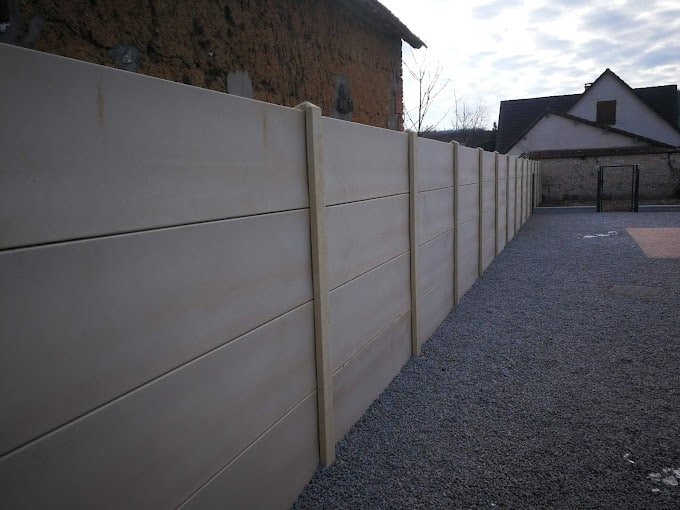 Clôture sur mesure en béton sans joints apparents, érigée le long d'un chemin en gravier avec des bâtiments en arrière-plan.