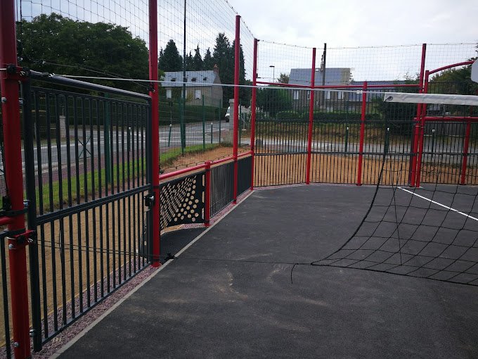 Intérieur d'un terrain multisports avec clôtures rouge et filet, et panneau de basket au fond.